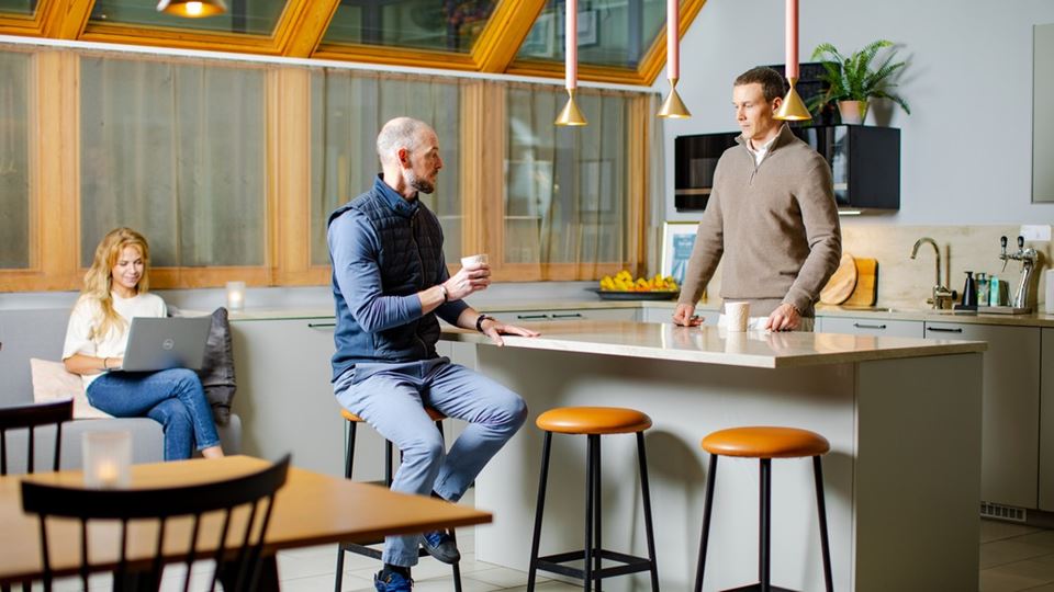 två personer står i ett kontoskök och pratar. en tredje sitter i en soffa bakom och tittar på datorn.