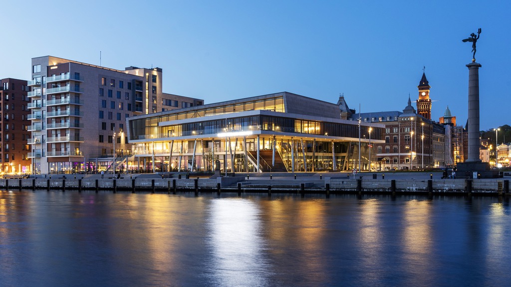 Bilden är tagen på kvällen och visar Perrongen 1 som lyser upp bilden med lamporna. Bilden är tagen från havet och man ser fastigheten med staden i bakgrunden.