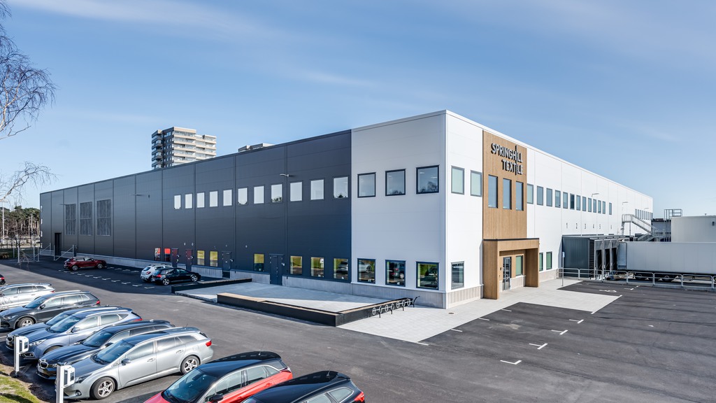 Bild tagen en solig dag med blå himmel. Visar byggnaden med vit och grå fasad, samt parkeringsmöjligheter för både cykel och bil utanför. Spring Hills logga över ingången och ett höghus i bakgrunden