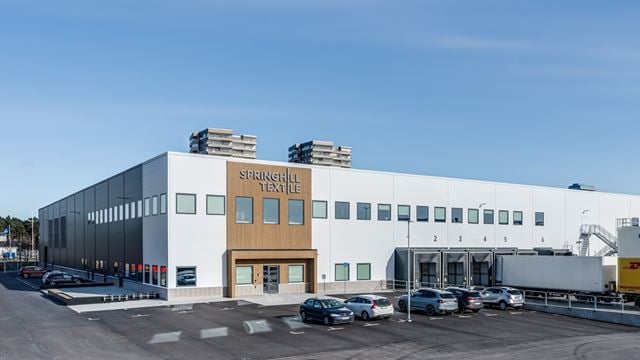 Bilden visar SpringHills nya lokal och dess lastbryggor. Utanför står en lastbil från DHL och några parkerade bilar. Det är en ljus bild med blå himmel. Fasaden är vit och vid ingången gjord av trä.