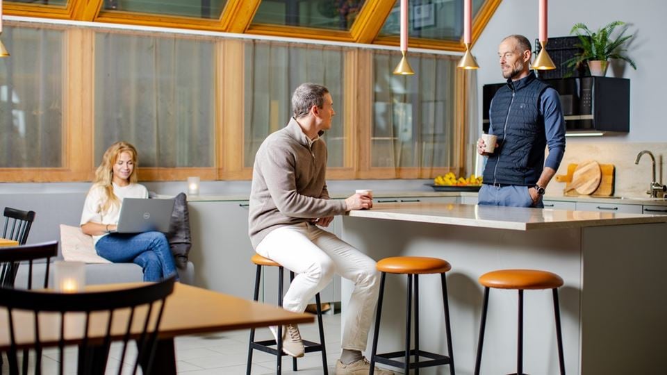 två personer står i ett kontoskök och pratar. en tredje sitter i en soffa bakom och tittar på datorn.