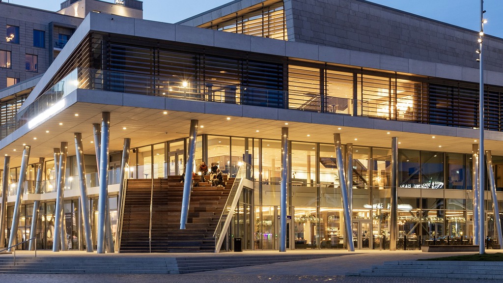 Visar huset på kvällen som lyser upp med lampor i ett varmt ljus. Man ser trappan som leder upp till ndra våningen. Det sitter människor i trappan