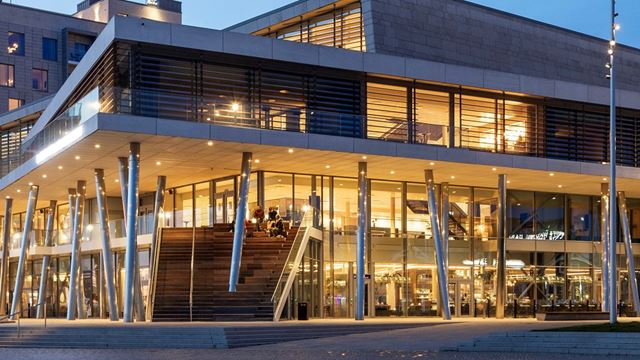 Visar huset på kvällen som lyser upp med lampor i ett varmt ljus. Man ser trappan som leder upp till ndra våningen. Det sitter människor i trappan