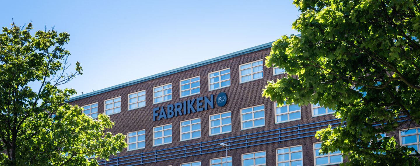 En mör tegelfasad med gröna träd i förgrunden. Skylt "Fabriken" på fasaden.