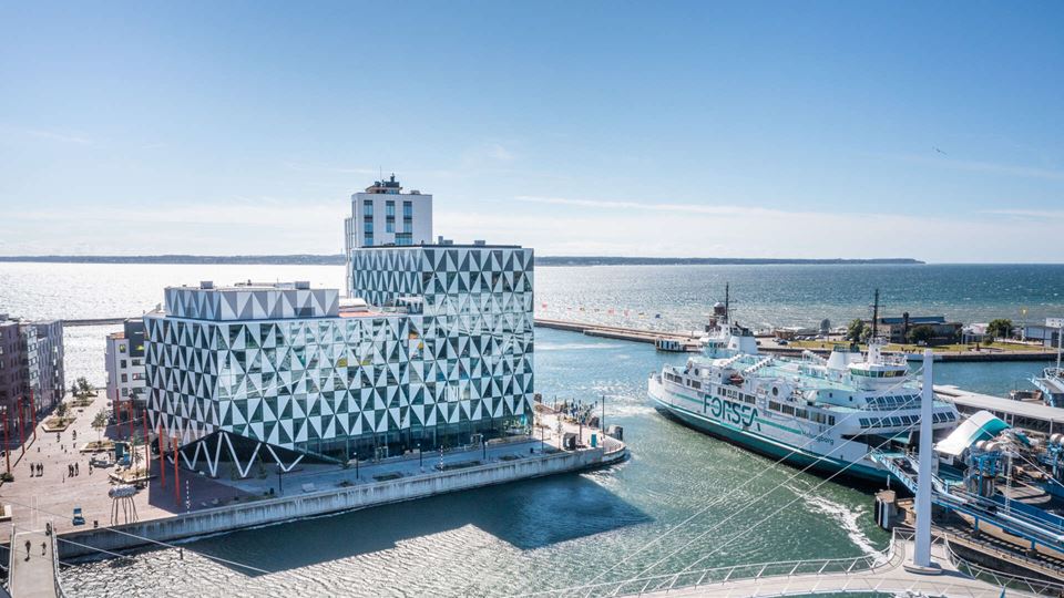 Fastigheten Prisma med en vackert mönstrad fasad, ligger i Oceanhamnen vid utloppet, en färja passerar förbi. En gång- och cykelbro svänger mot kajen. 